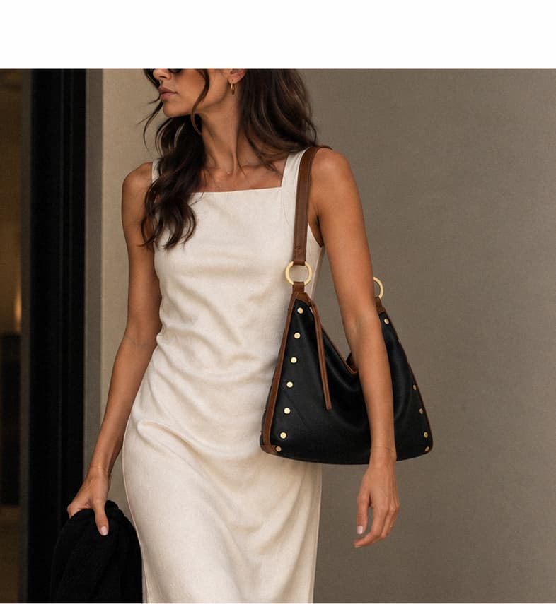 Model in a white dress carrying a black leather shoulder bag with brown trim and gold rivet detailing.