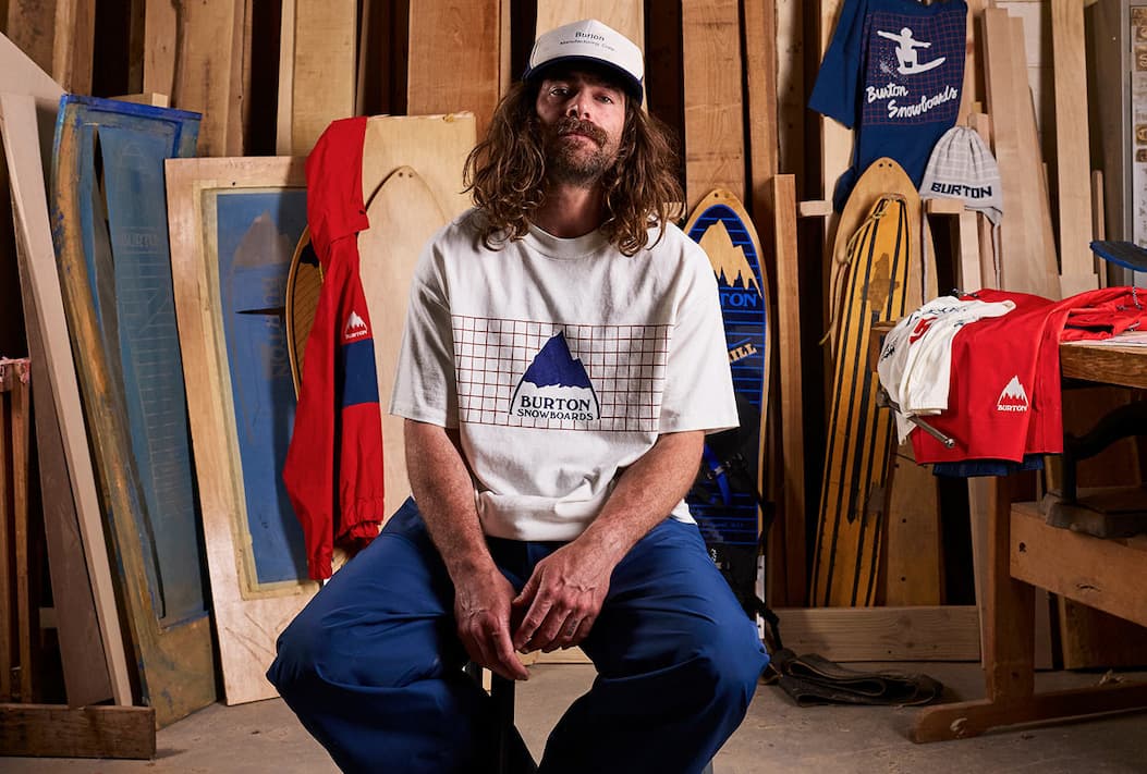 Man in a Burton Snowboards t-shirt and cap sitting in a wood workshop surrounded by vintage snowboards and gear.