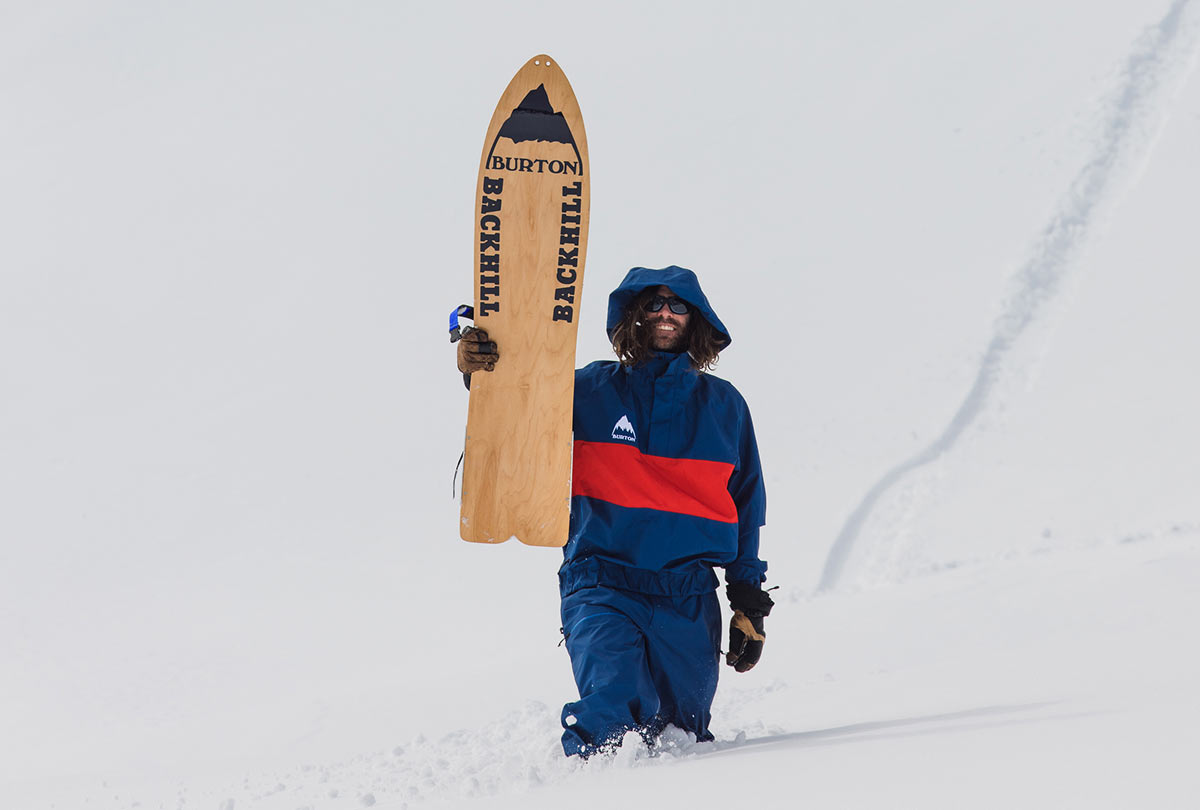 Man in retro blue and red jacket walking through snow, holding a wooden Burton Backhill snowboard.