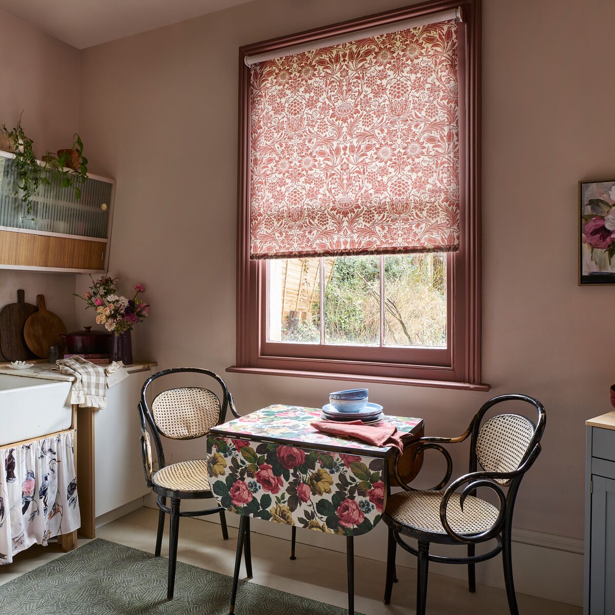 Floral Roman blind framed by a wooden window, filtering daylight into a cosy dining nook with matching botanical fabrics.