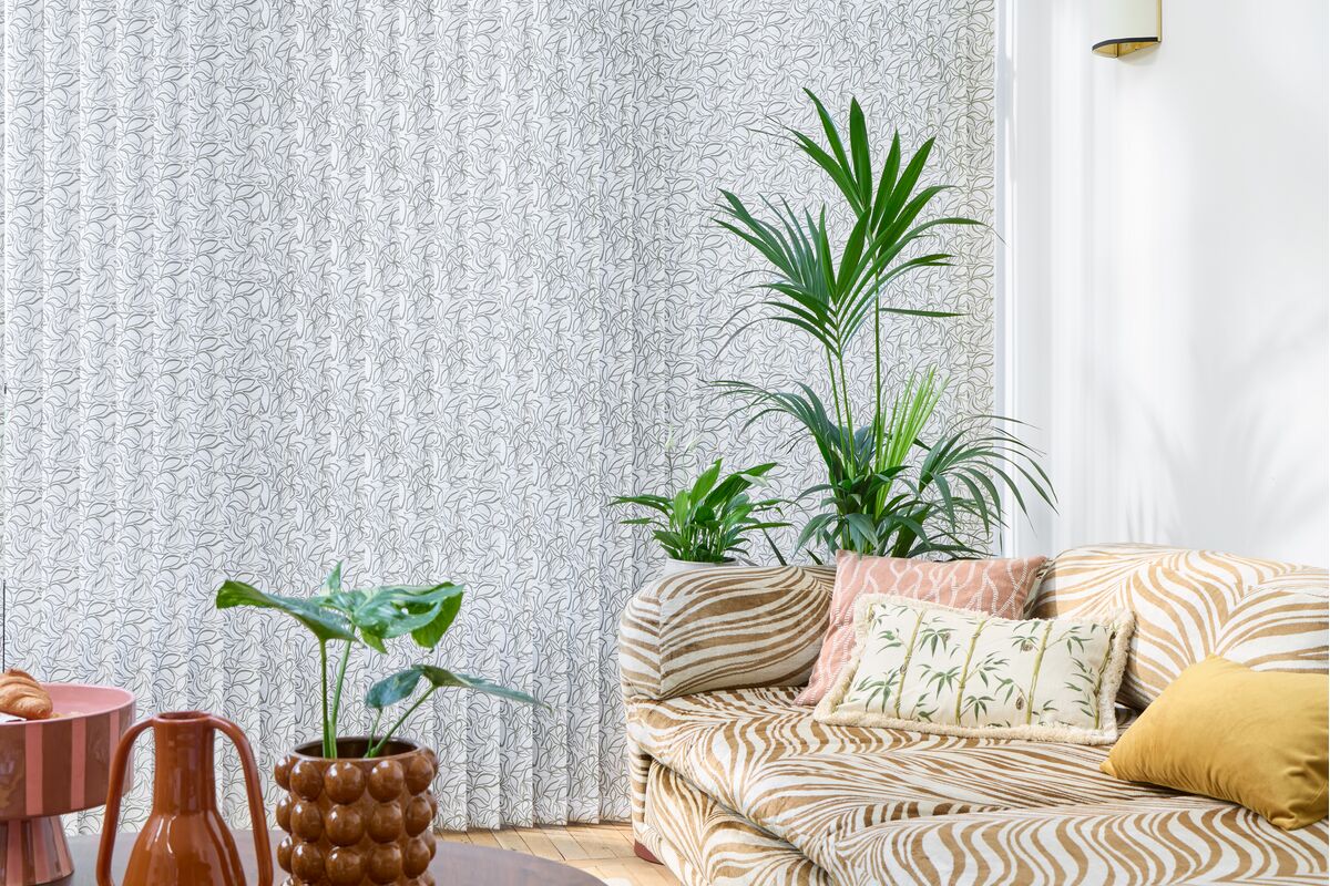Floor-to-ceiling vertical blinds with a delicate leaf pattern create a soft backdrop in a bright living space with plants and a striped sofa.