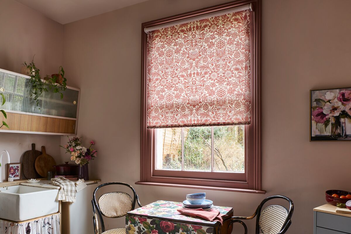 Floral Roman blind framed by a wooden window, filtering daylight into a cosy dining nook with matching botanical fabrics.