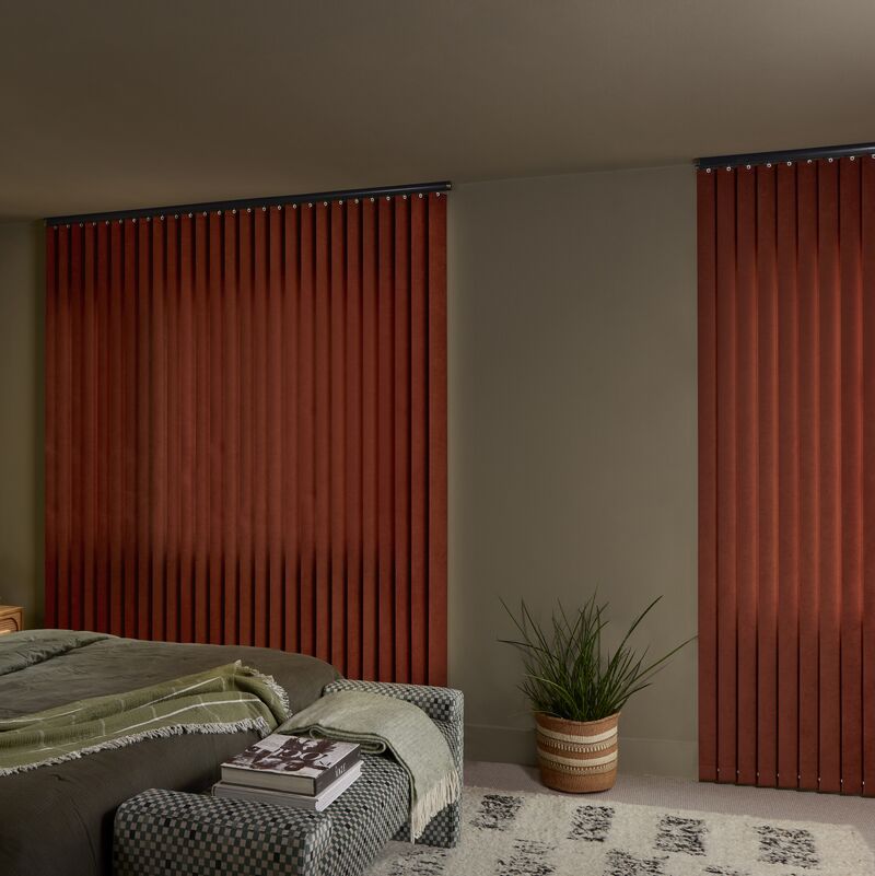 Rust-coloured Vertical blinds cover two bedroom windows, complementing earthy bedding, a patterned bench and potted plant.