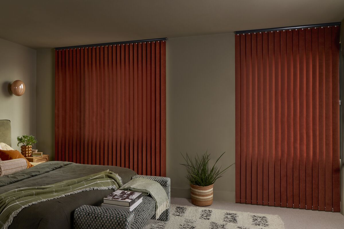 Rust-coloured Vertical blinds cover two bedroom windows, complementing earthy bedding, a patterned bench and potted plant.
