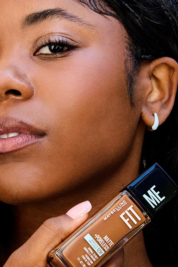 Close-up of a model with a brown skin tone wearing Maybelline Fit Me Matte + Poreless Foundation and holding the bottle of her matching shade next to her chin, highlighting a blurred matte complexion with visibly smooth-looking skin.