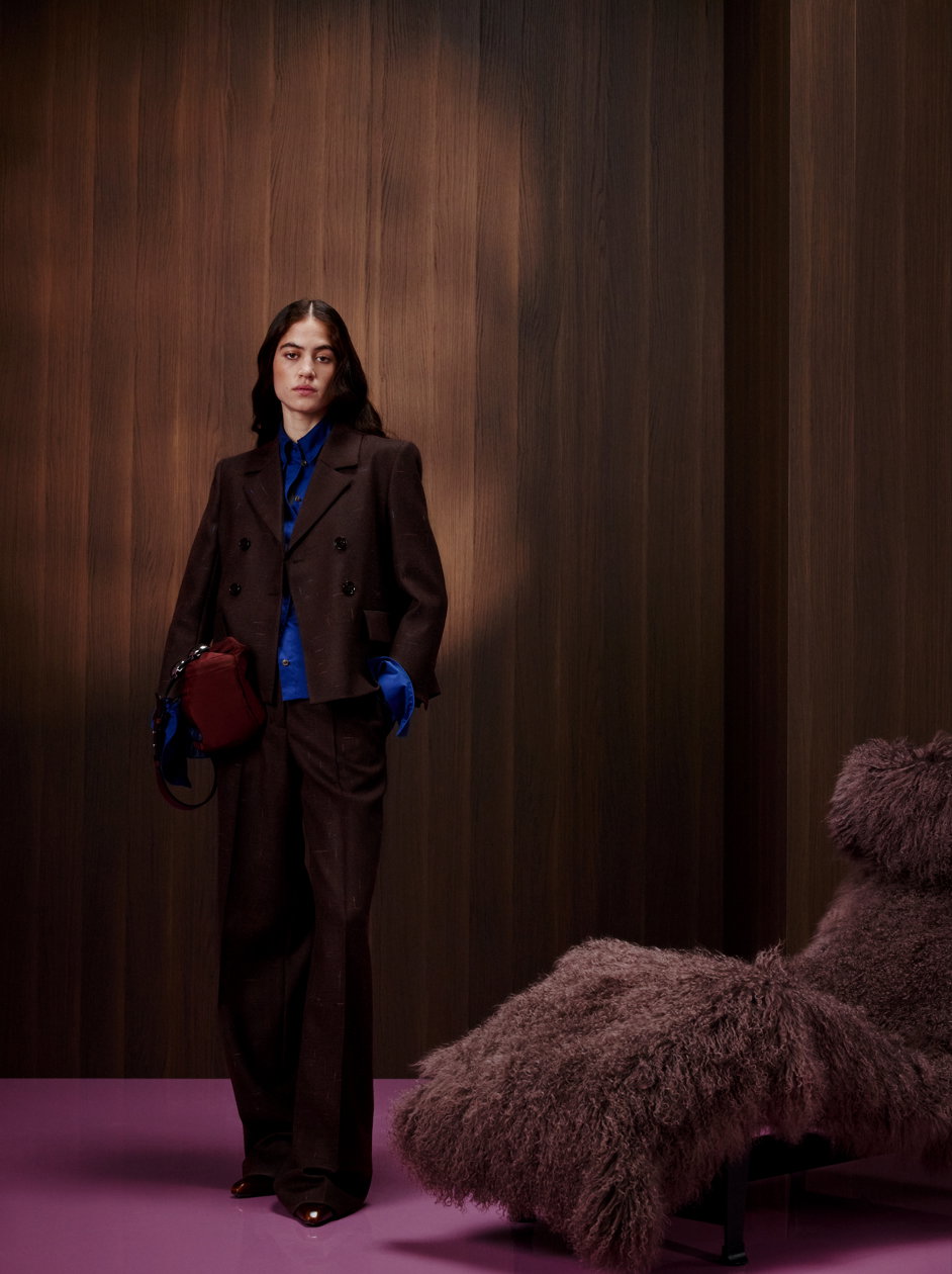 Ein Model von DOROTHEE SCHUMACHER steht vor einer dunklen Holzwand auf einem glänzenden lila Boden. Sie trägt einen eleganten, dunkelbraunen Hosenanzug mit zweireihigem Blazer, kombiniert mit einer leuchtend königsblauen Bluse und einer bordeauxroten Handtasche, neben einem flauschigen Lounge-Sessel.