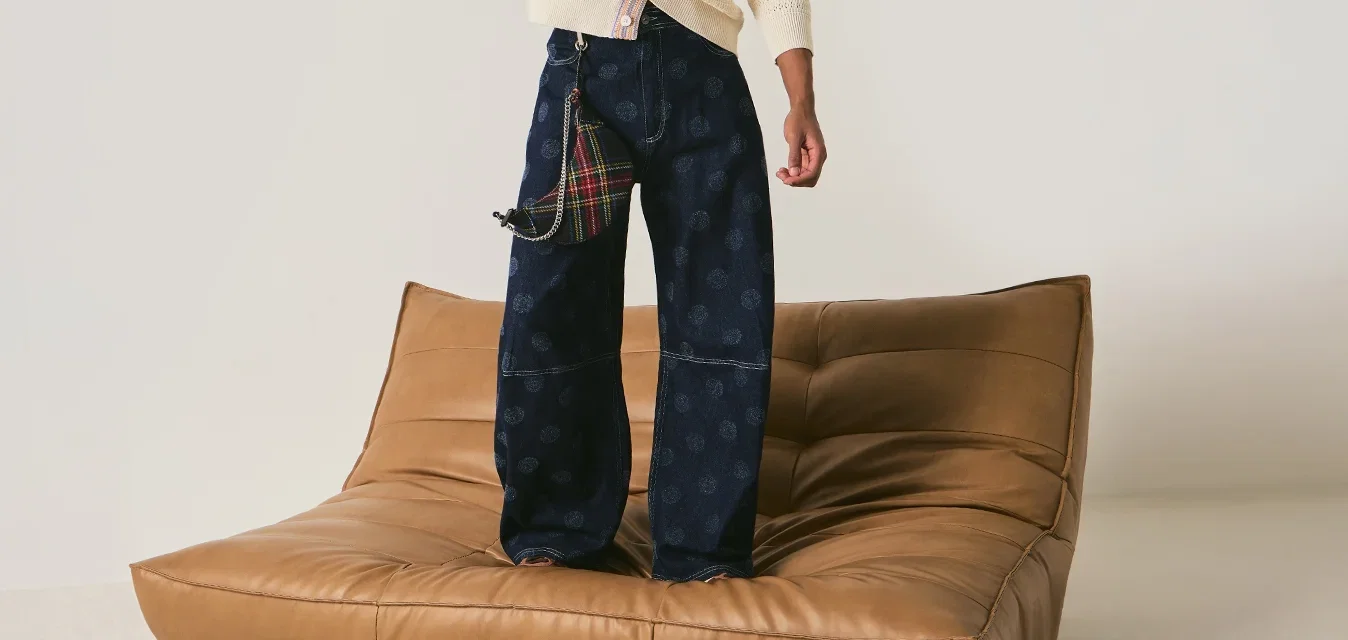 Jonge man die op een lage tan leren bank staat, gekleed in een crème cardigan en wijde marineblauwe broek, terwijl hij zijn hoed aanraakt in een minimalistische witte kamer.