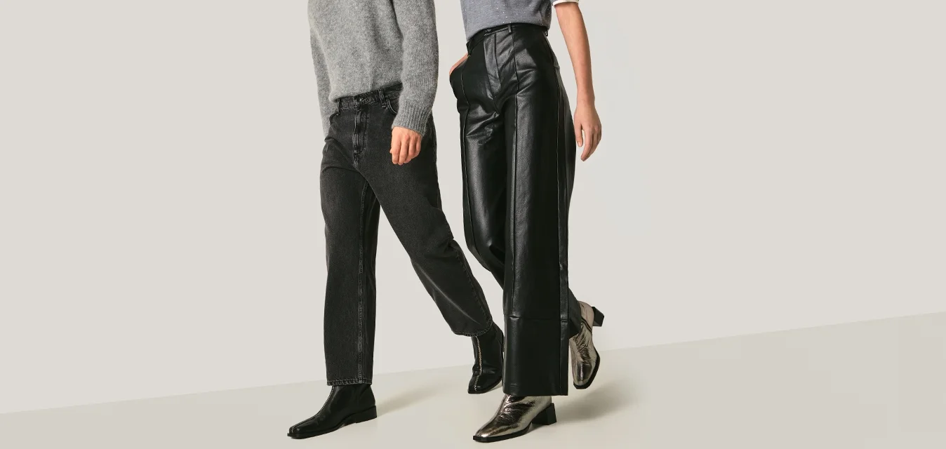 Man en vrouw lopen naast elkaar, gekleed in grijze bovenkleding, zwarte broeken en enkellaarsjes, tegen een effen lichte achtergrond.
