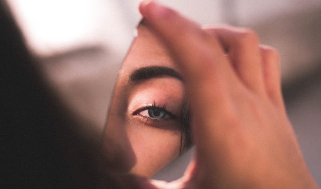 A person holds a small mirror shard reflecting a close-up of one eye and part of an eyebrow.