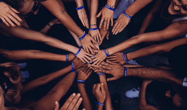 A group of people standing in a circle with their hands extended towards the center, wearing blue bracelets.