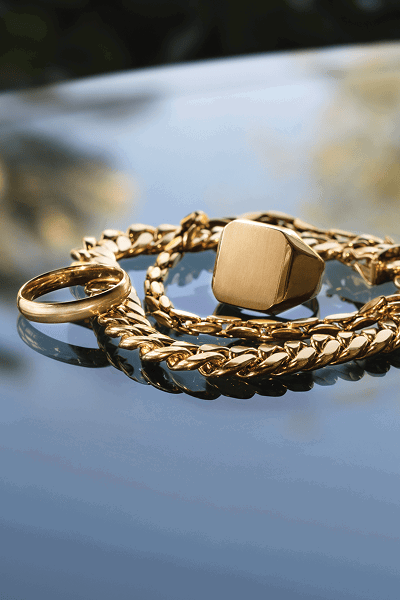 Close up of a men’s yellow gold chain and rings on reflective surface.