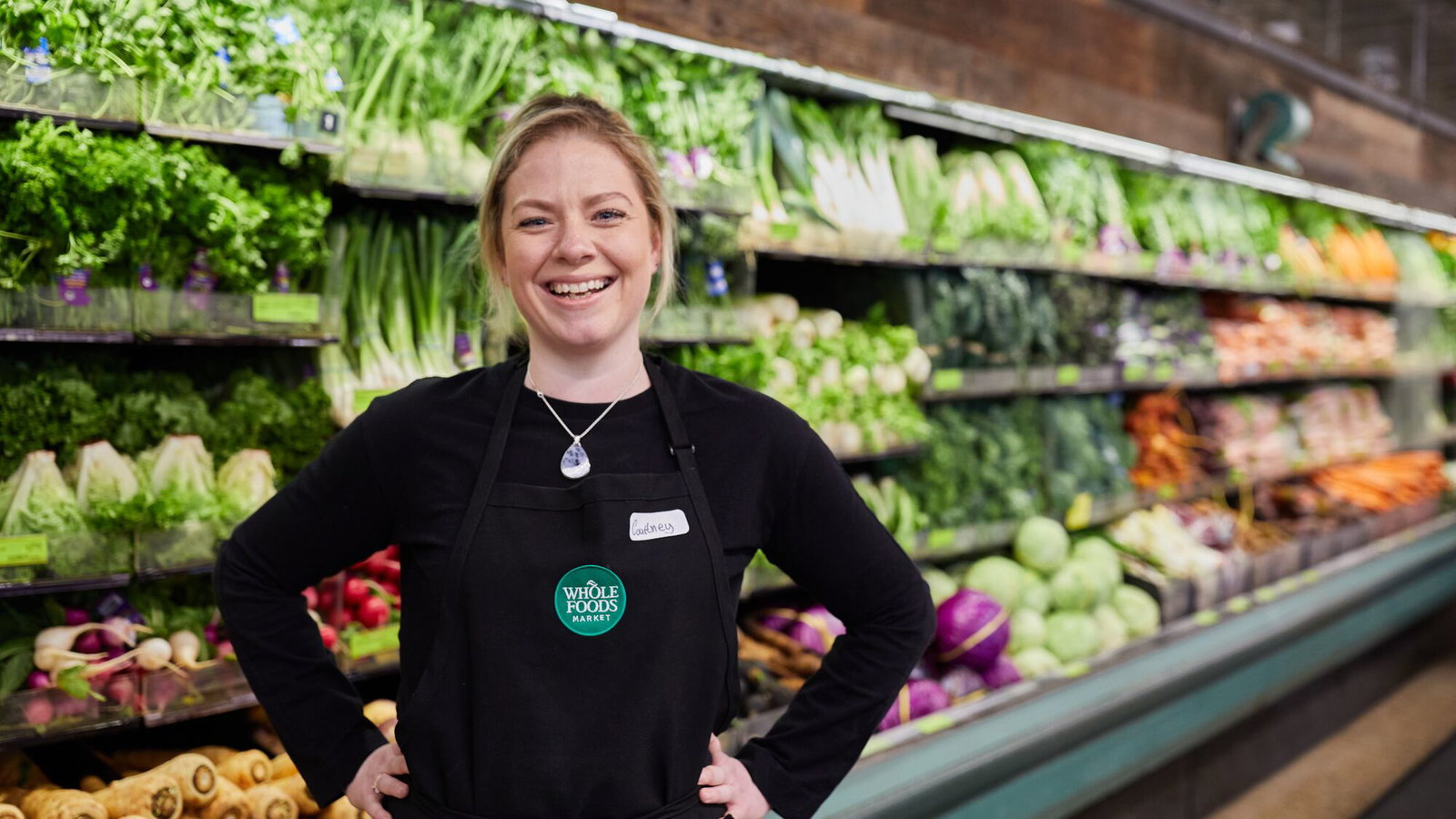 Smiling Whole Foods Market team member stands before a vibrant refrigerated display of fresh vegetables.