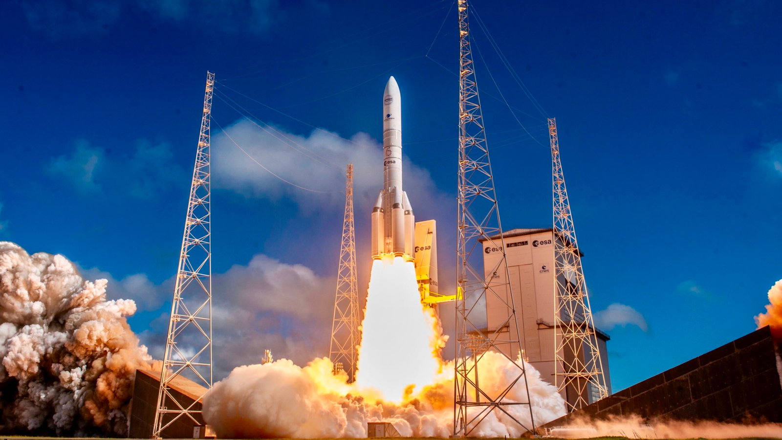 A rocket lifts off from its launch pad, framed by towers, fire, and billowing smoke.