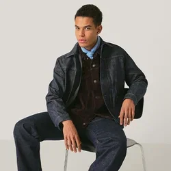 Jonge man die een donker gestructureerd jack, blauw overhemd, corduroy gilet, spijkerbroek en zwarte laarzen draagt, zittend op een stoel tegen een effen achtergrond.