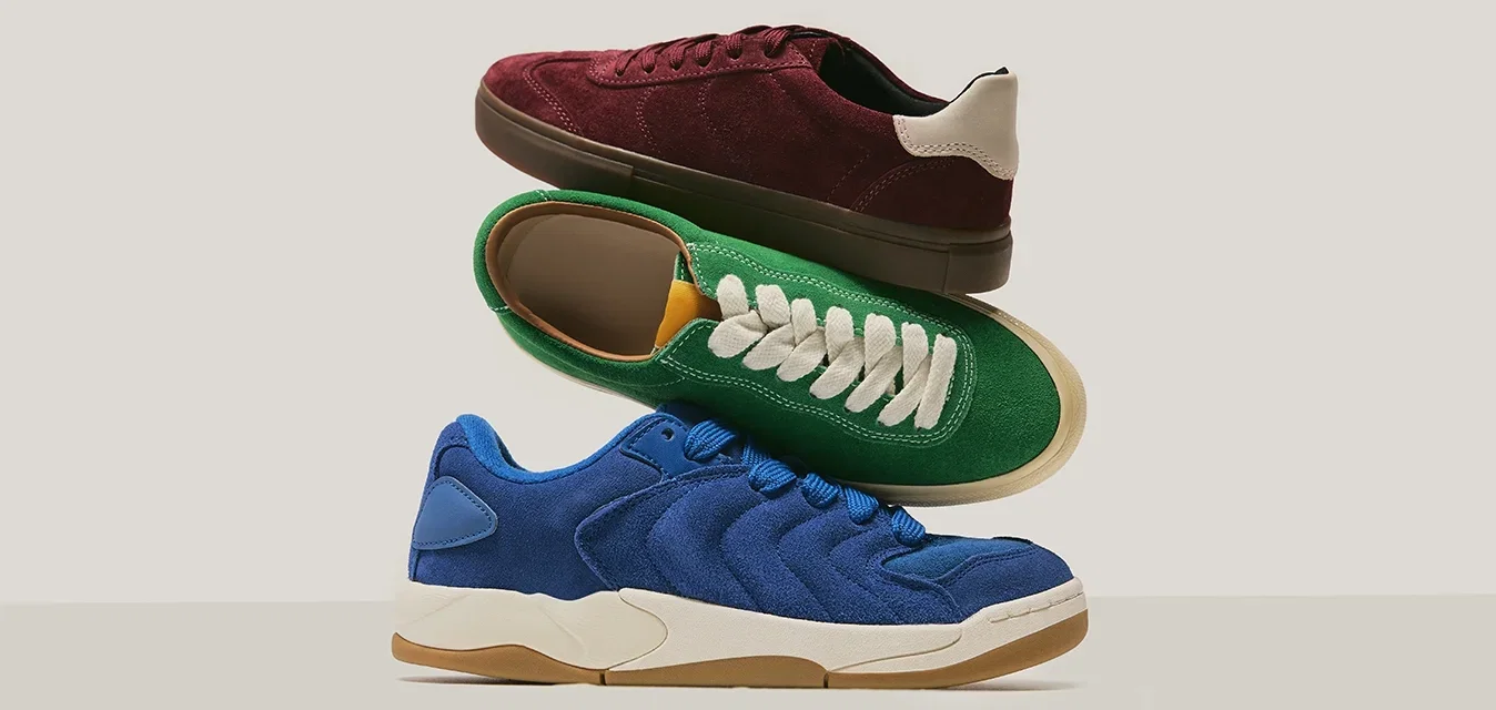 Drie op elkaar gestapelde suède sneakers in blauw, groen met witte veters en bordeauxrood met beige hiel-detail op een neutrale achtergrond.
