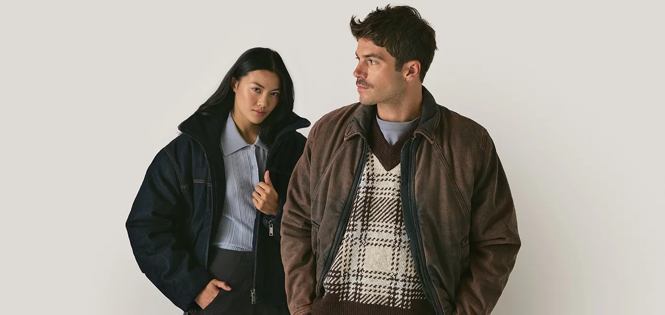 Twee personen dragen jassen: de een in een donkere denim puffer en de ander in een bruin leren jack over een gestructureerde trui. Beide hebben een slim fit.