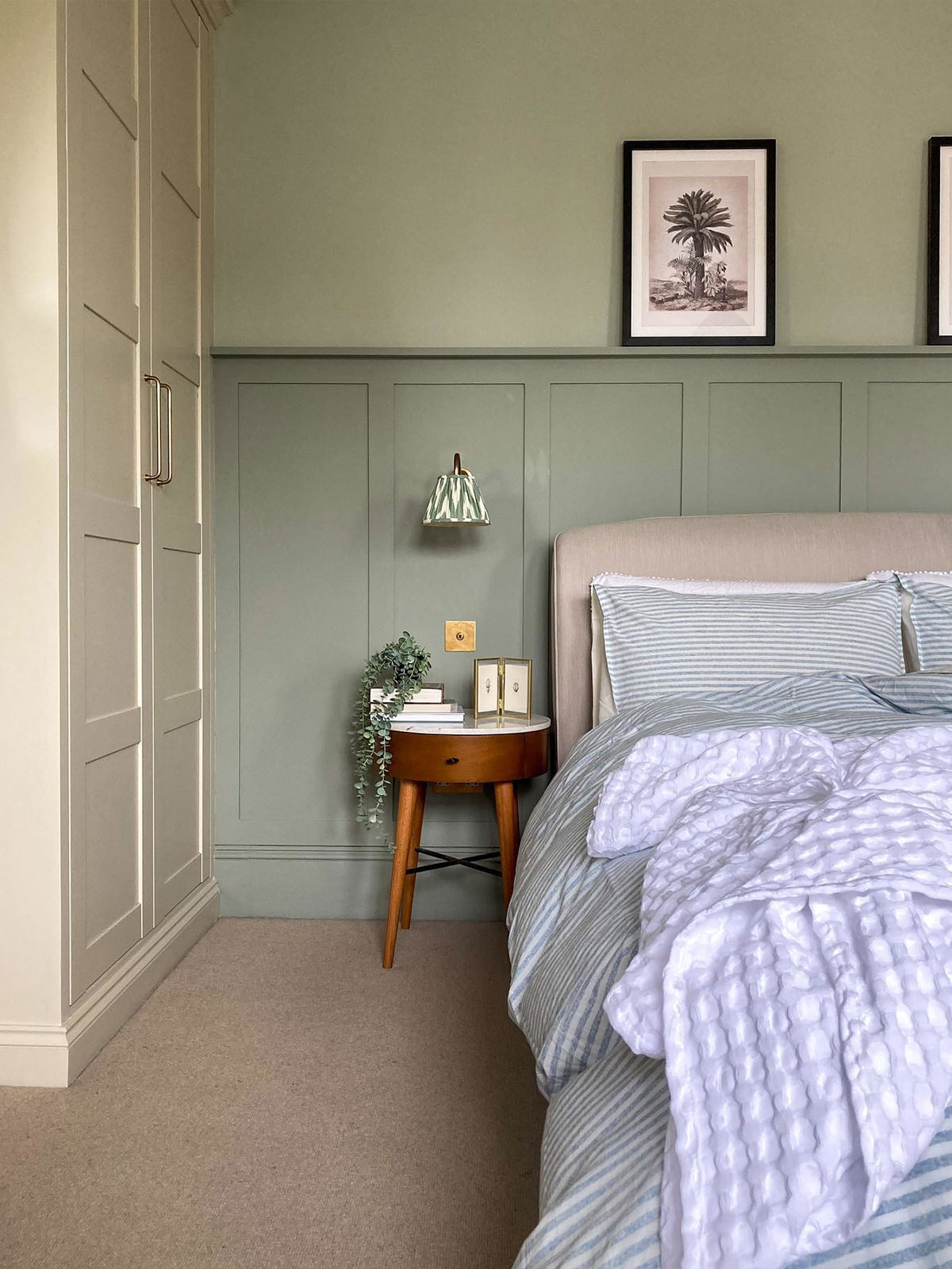 A calm, classic bedroom. The walls are painted in a light grey with a darker, blue-grey panelling on the bottom half. A double bed with plump, striped bedding is in front of the panel. The room feels peaceful and reassuring, ready for a good night’s sleep.