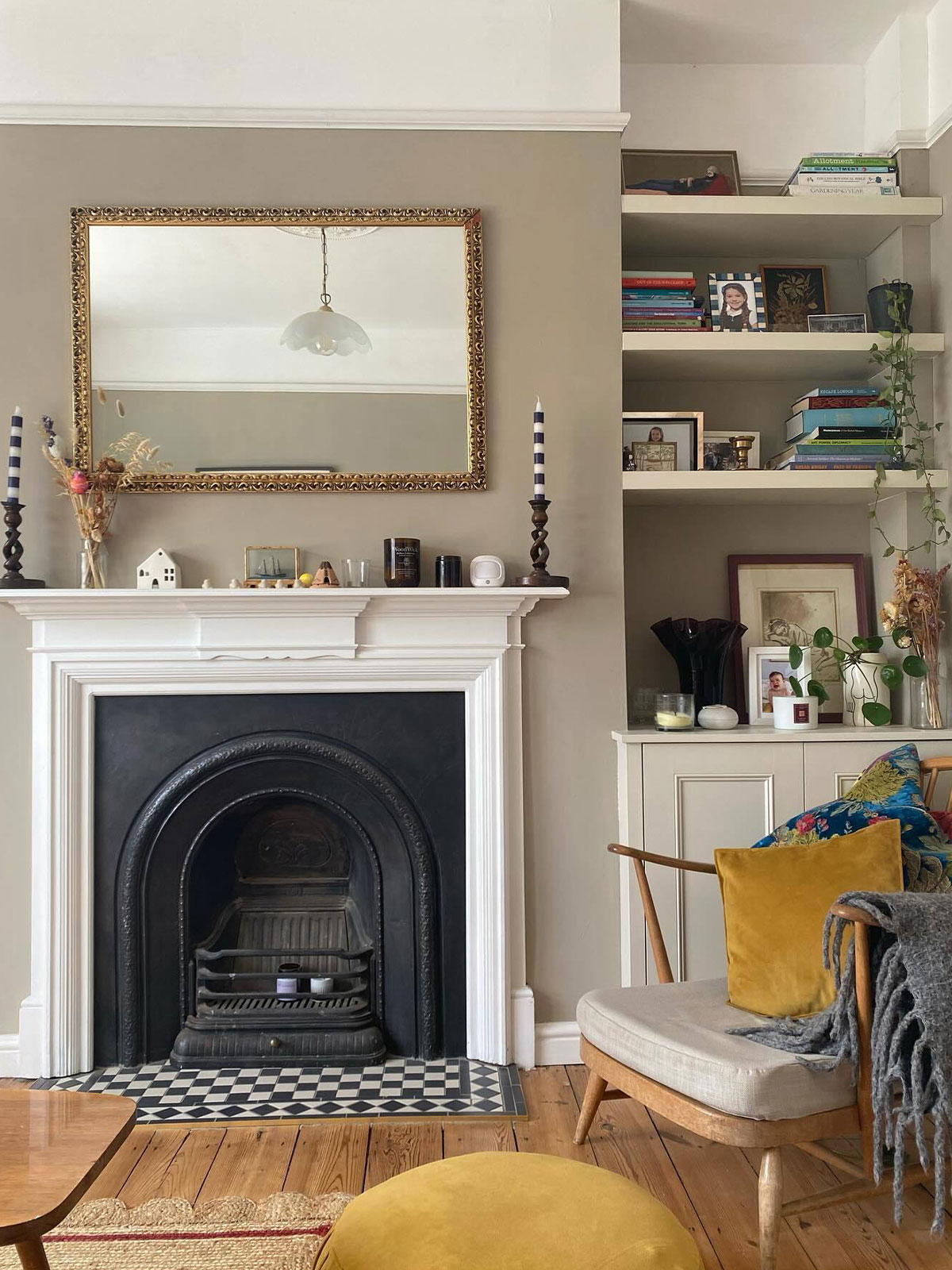 Inviting living room with period features, including a picture rail and black, cast iron fire place and black and white tile hearth. The furniture is wooden, mid-century modern, which adds a touch of modernity to the traditional space. There are books, plants, photos and ornaments dotted around the space, making it feel lived-in.