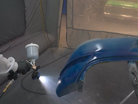 Spray gun applying blue paint to a curved automotive body panel inside a paint booth.
