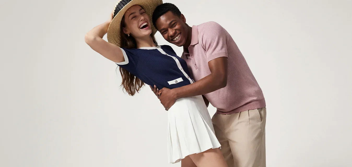 Jonge man en vrouw lachen en omhelzen elkaar, vrouw draagt hoed, marineblauw topje en witte rok, man draagt roze blouse en beige broek op witte achtergrond.