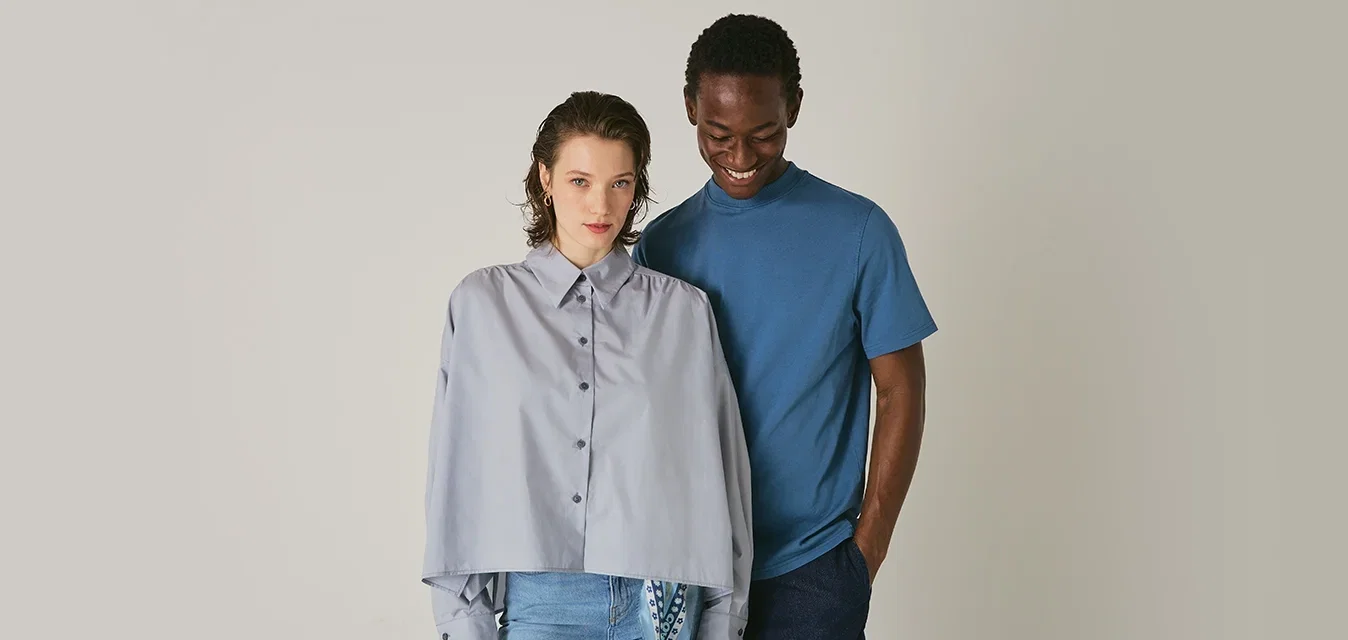 Lichtgrijs overhemd met oversized mouwen, gecombineerd met een blauwe t-shirt; beide hebben casual stijlen tegen een neutrale achtergrond.