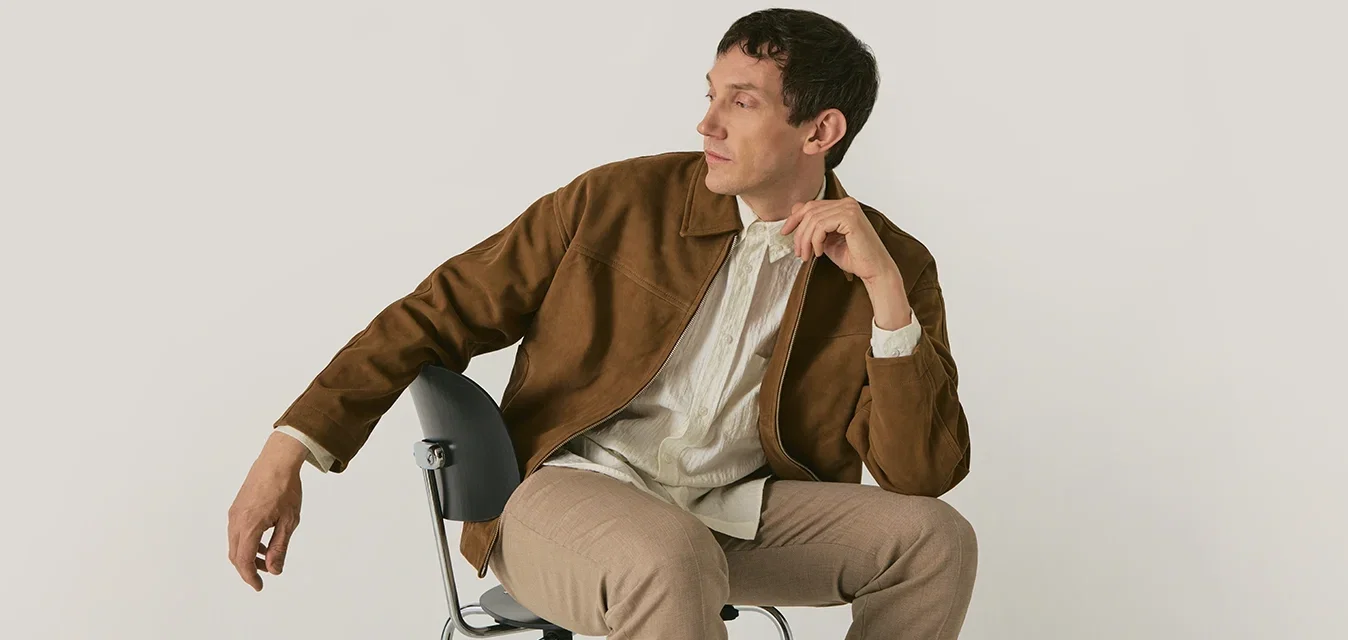 Man in een bruine jas en beige broek zittend zijwaarts op een zwarte stoel, wegkijkend met zijn hand dicht bij zijn gezicht tegen een effen achtergrond.