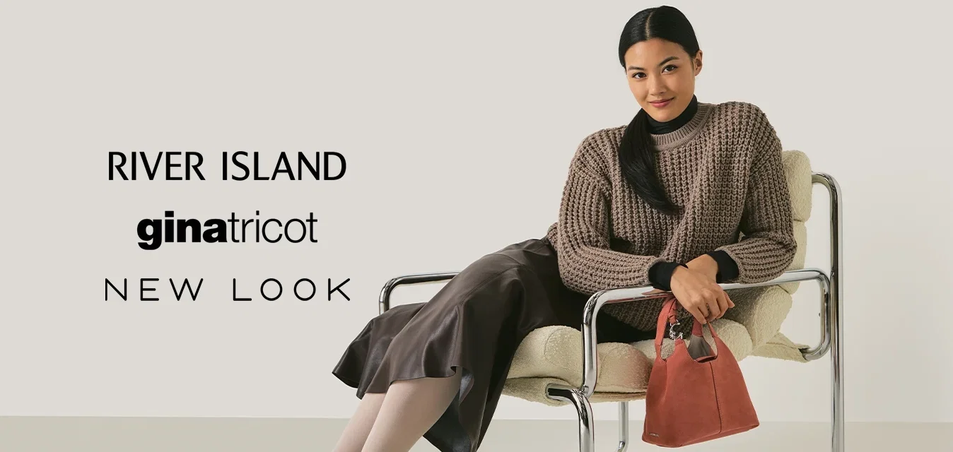 Vrouw in een bruine gebreide trui en leren rok, zittend op een moderne chrome en stoffen stoel, met een kleine rode handtas in haar hand, lachend naar de camera.