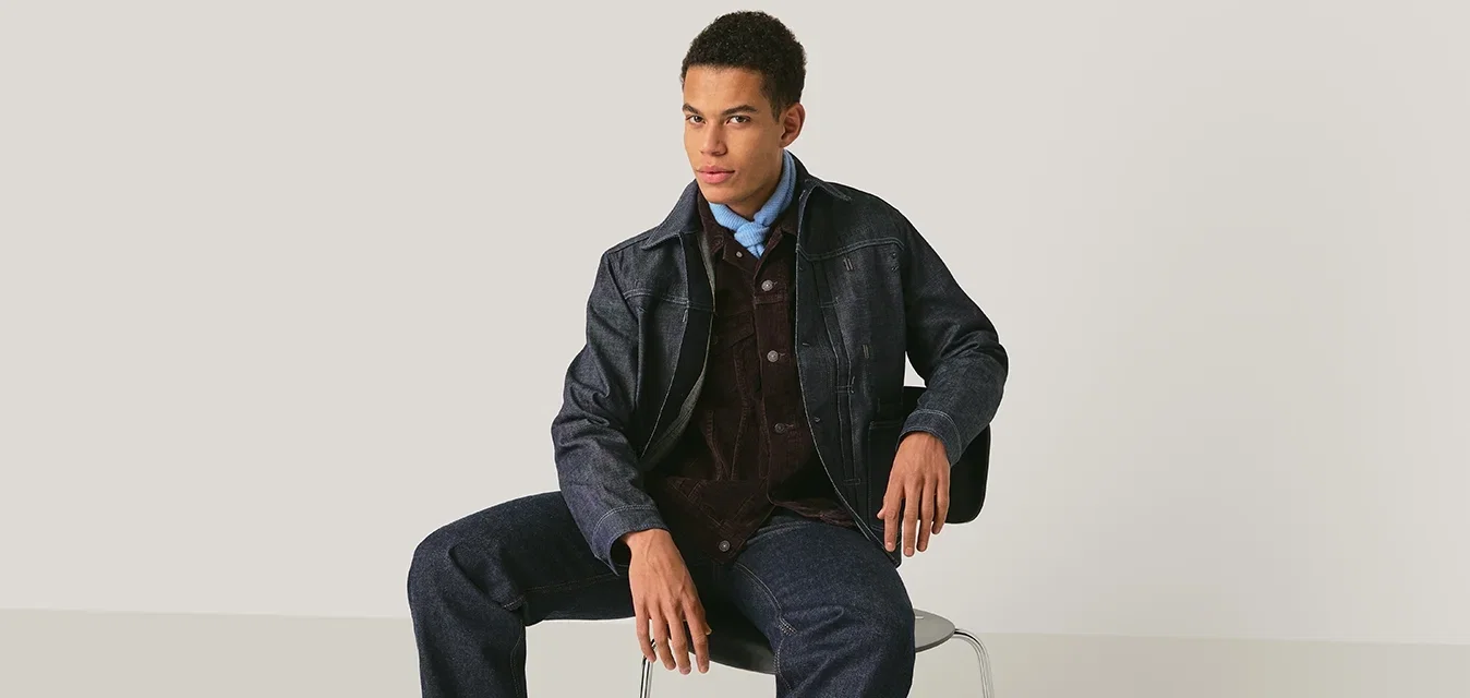 Jonge man die een donker gestructureerd jack, blauw overhemd, corduroy gilet, spijkerbroek en zwarte laarzen draagt, zittend op een stoel tegen een effen achtergrond.