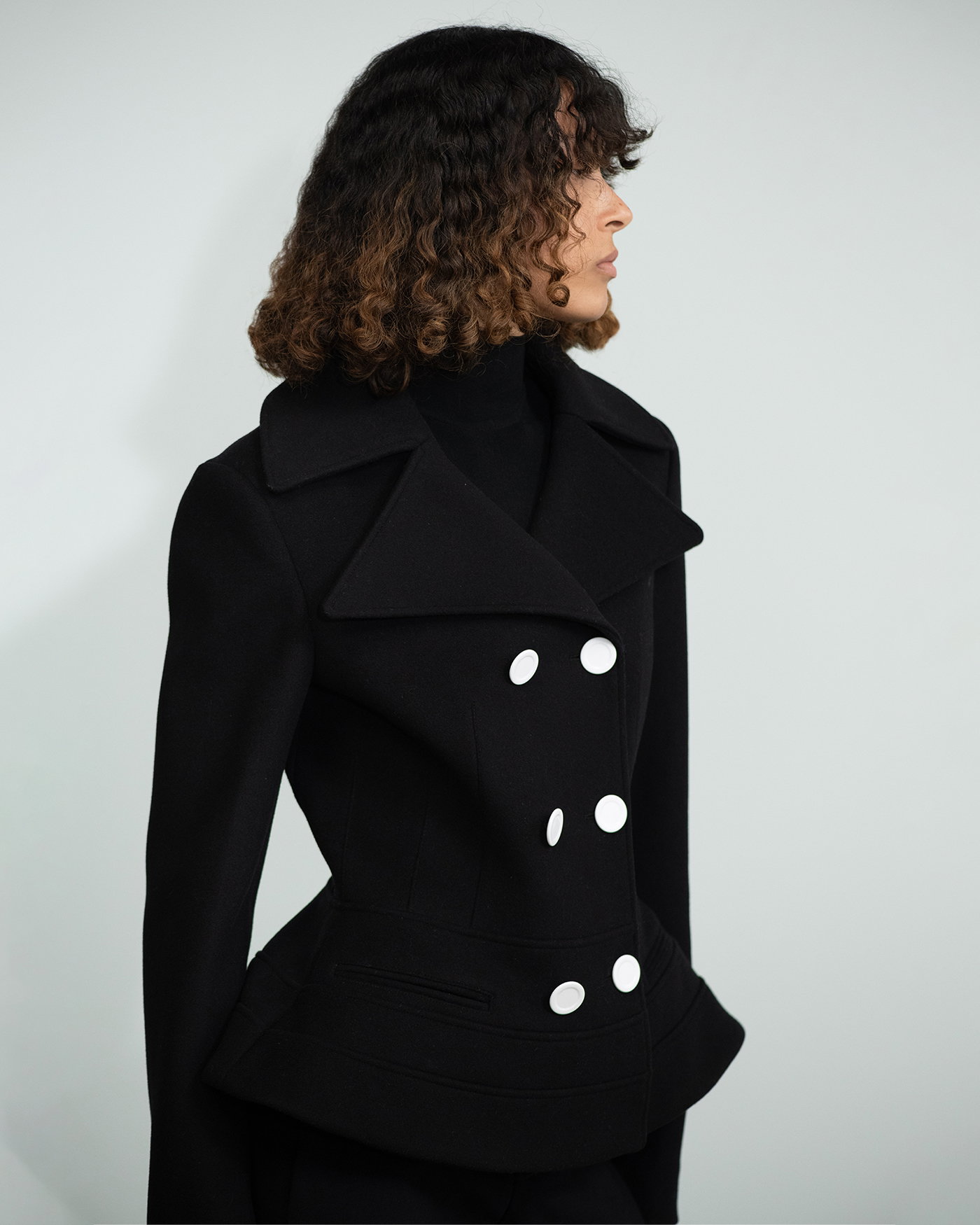 Cropped Image of model standing in front of White Wall wearing the Romane Jacket in Compact Melton Wool in Black