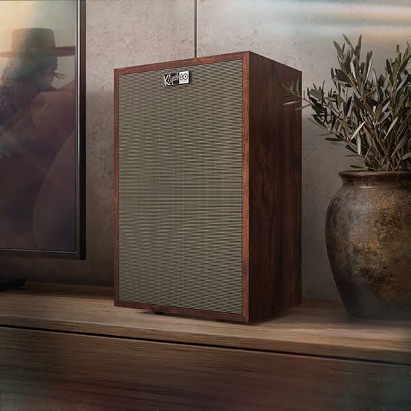 Curated living room with a Klipsch Rebellion speakers situated on a wooden cabinet, between a flatscreen tv and a potted plant.