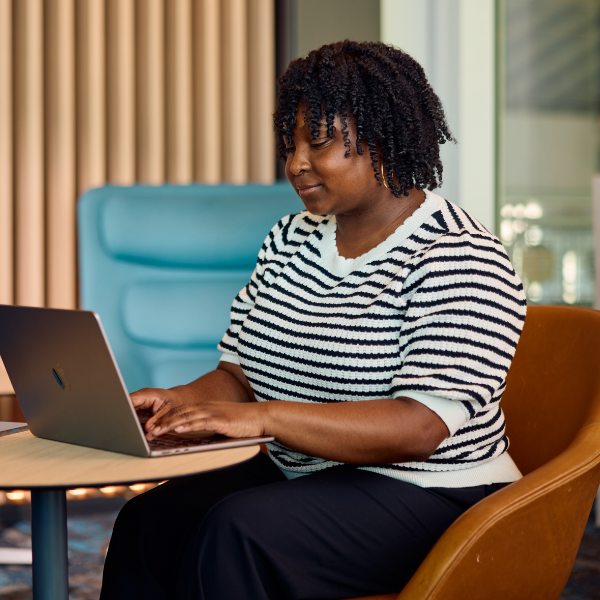  A woman sits at a small round table working on a laptop computer. She is in a contemporary office or coworking space with modern chairs and vertical acoustic panels in the background. The scene suggests focused work or remote collaboration in a comfortable, professional environment. Warm lighting and soft furnishings create a welcoming workplace atmosphere.