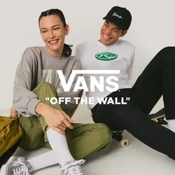 Twee jonge mensen zitten op de grond in casual kleding, één met een skateboard en een gele rugzak, ze lachen en zitten dicht bij elkaar.