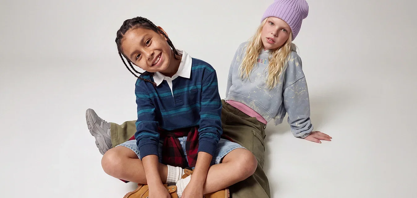 Twee kinderen zitten op de vloer, één glimlacht in een gestreepte trui en laarzen, de ander in een muts en casual kleding leunt achterover.