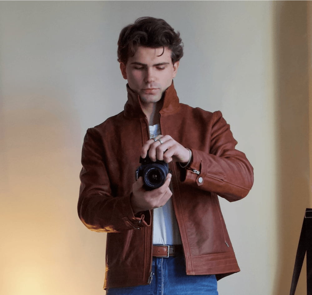 Man in a brown leather jacket holding a camera, standing indoors against a plain wall.