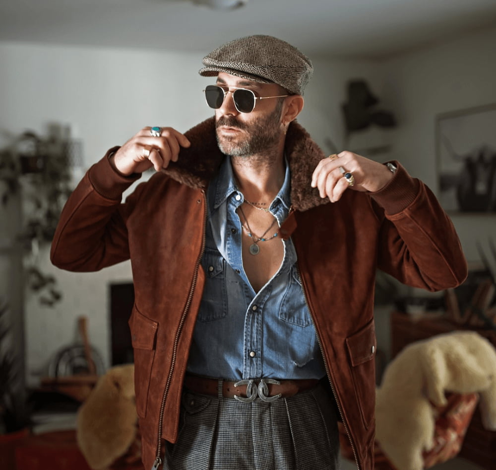 Man in sunglasses and a flat cap wearing a brown suede shearling-collar jacket over a chambray shirt, indoors.