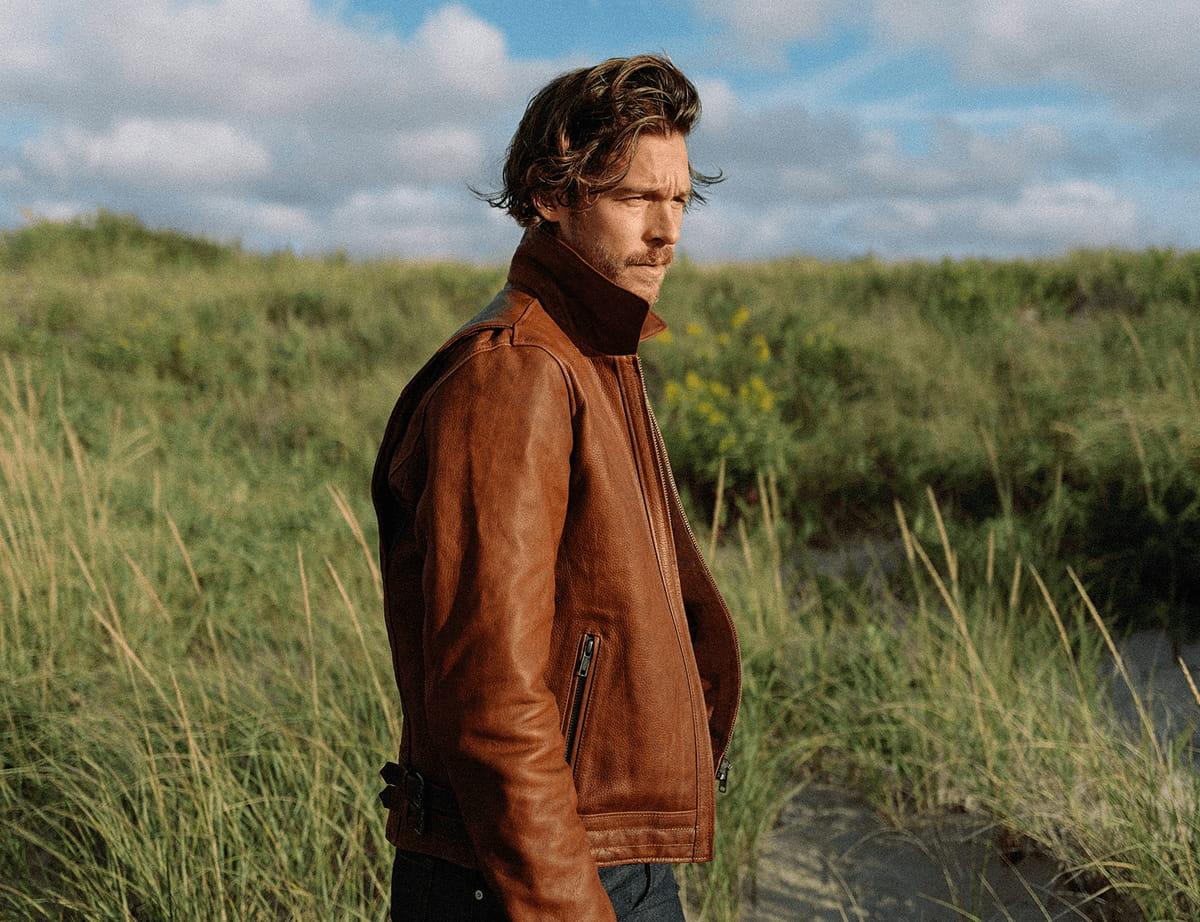 Man in a brown leather moto jacket standing in tall dune grass under a partly cloudy sky.