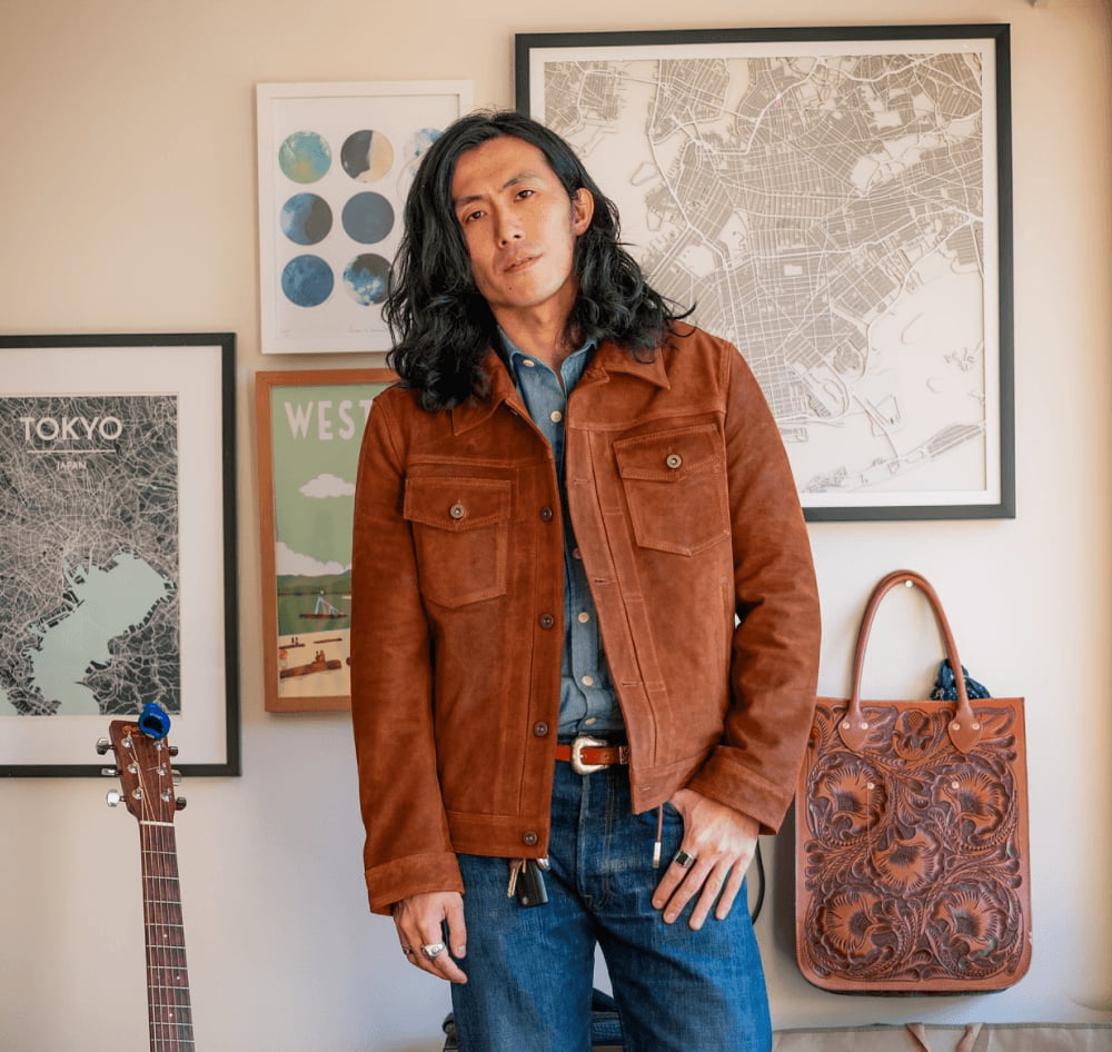 Man in a brown suede jacket standing in front of framed city maps and a tooled leather tote bag.
