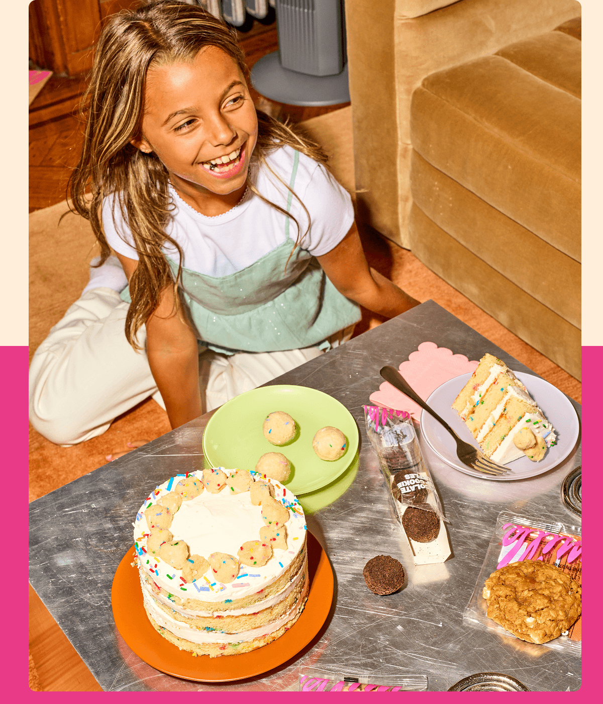 Smiling girl seated beside a table with a Birthday Cake, a slice of cake, Birthday Cake Truffles, and a packaged cookie.