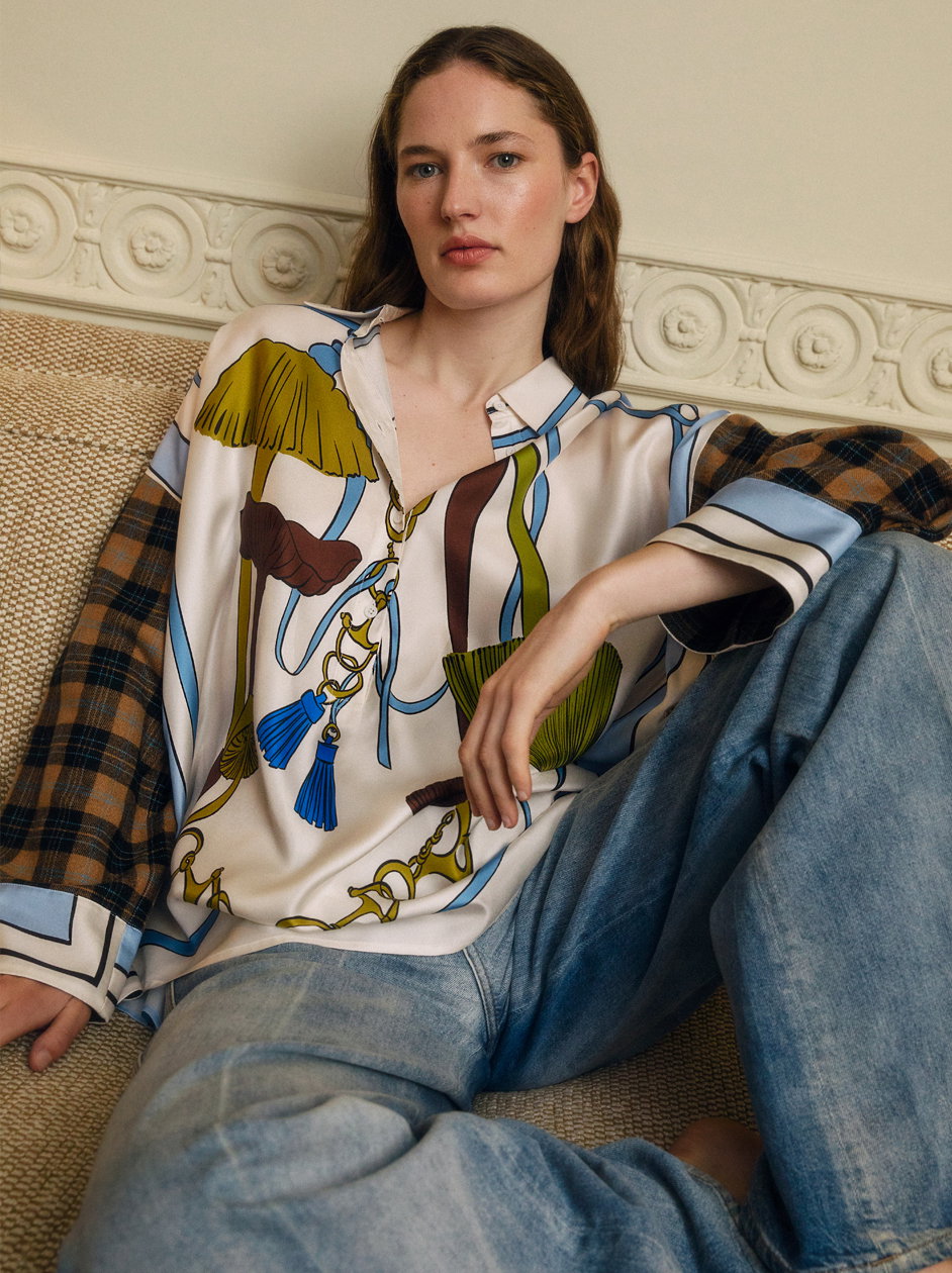 Ein Model von DOROTHEE SCHUMACHER sitzt entspannt auf einem beigen Sofa. Sie präsentiert eine fließende Seidenbluse im ausdrucksstarken Foulard-Print mit Quasten-Motiven und kontrastierenden karierten Ärmeln, lässig kombiniert mit einer weiten Jeans.