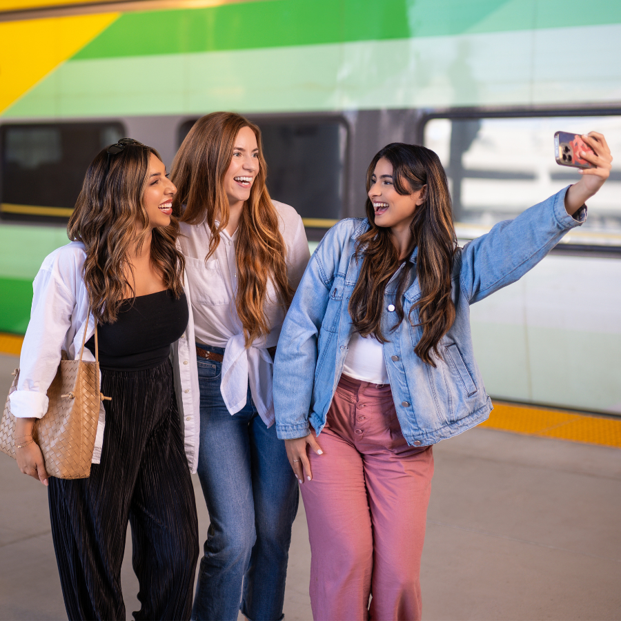 Three young women stand together on a train platform, smiling as one holds up a smartphone to take a selfie. A modern train with bold green and yellow stripes is visible in the background.