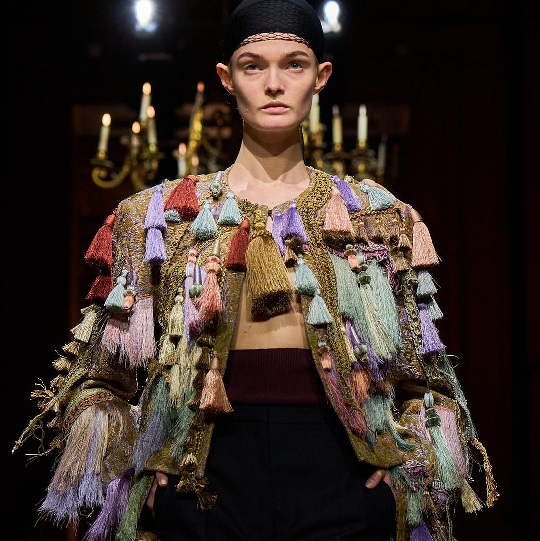 Model showcasing a colorful, tassel-adorned jacket paired with dark pants and a head covering on a runway. Model showcasing a colorful, tassel-adorned jacket paired with dark pants and a head covering on a runway.
