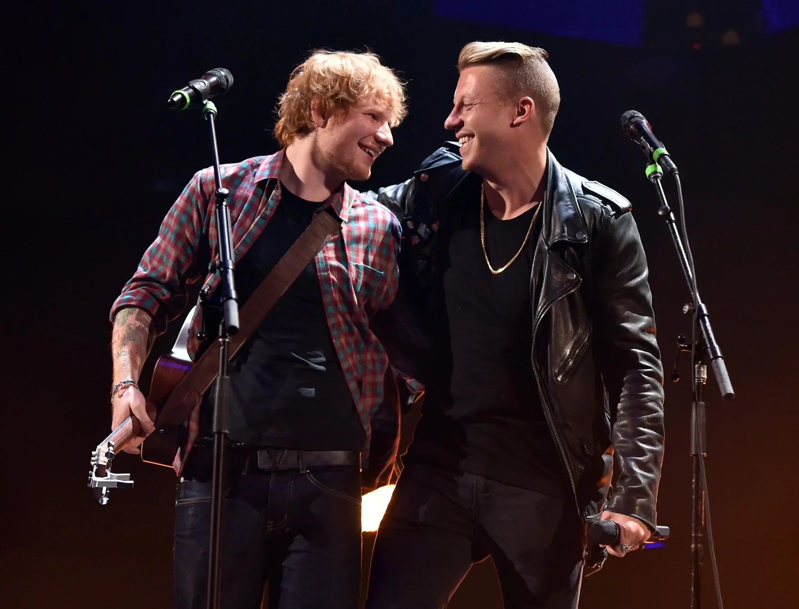 Ed Sheeran and Macklemore smile at each other, arms around each other, on stage with two microphones.