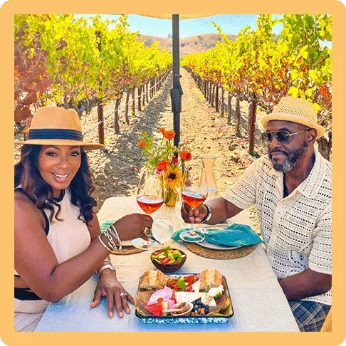Image Description: Couple seated at a table and toasting with two glasses of wine with rows of vineyards behind them.