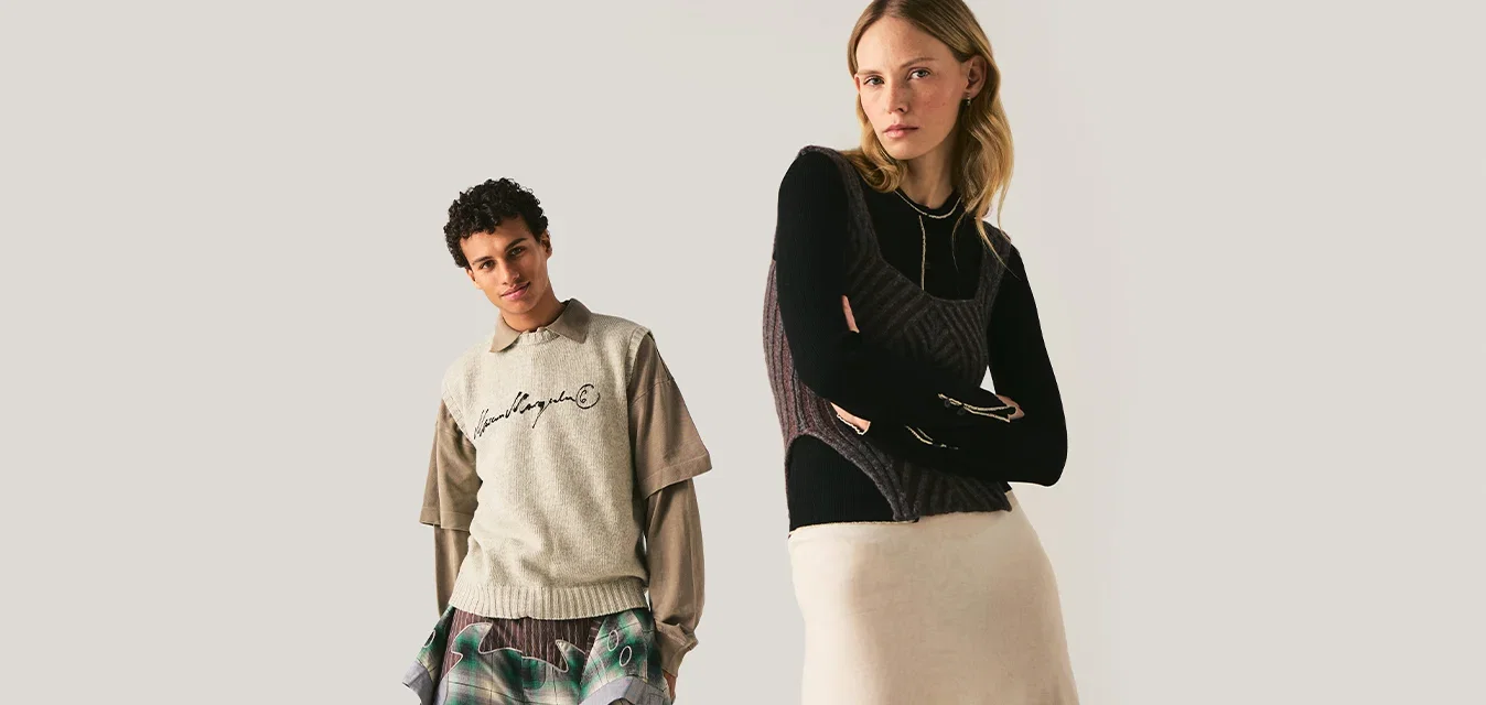 Een man en een vrouw staan tegen een effen achtergrond, gekleed in gelaagde, moderne casual kleding in ontspannen houdingen.