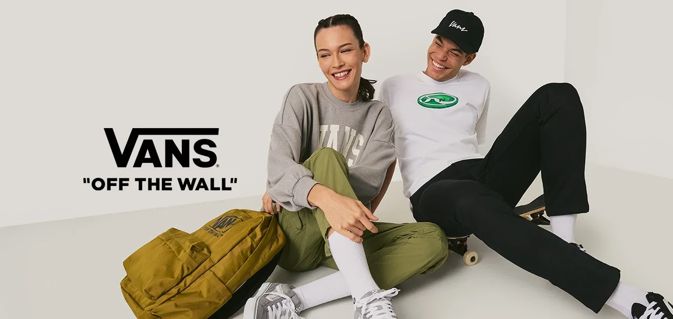 Twee jonge mensen zitten op de grond in casual kleding, één met een skateboard en een gele rugzak, ze lachen en zitten dicht bij elkaar.