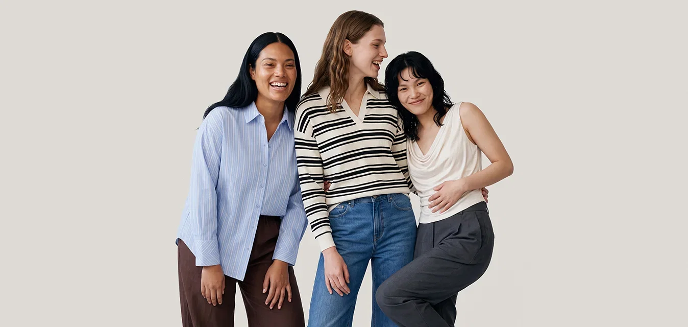 Drie vrouwen staan dicht bij elkaar, glimlachend en lachend, gekleed in casual kleding tegen een effen lichte achtergrond.