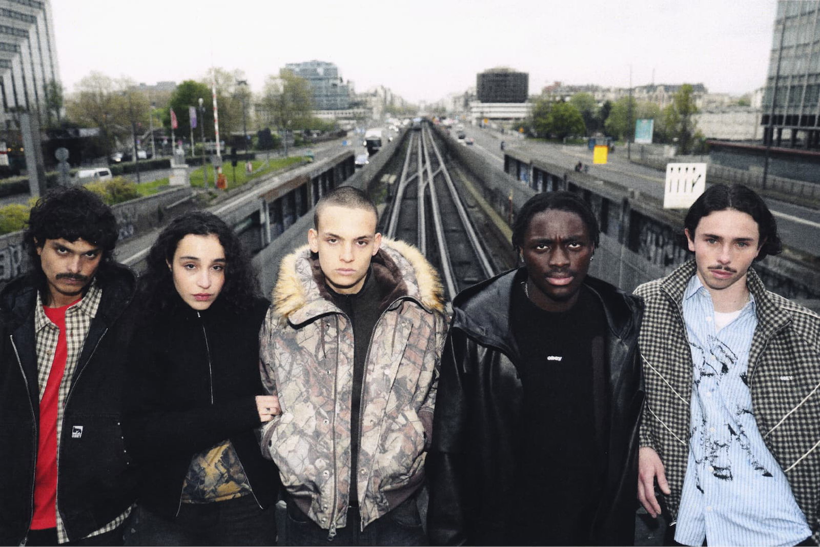 Five models standing side-by-side above train tracks in an urban setting, showcasing Obey jackets and coats including a black work jacket, camo hooded parka, dark leather jacket, and patterned overcoat.