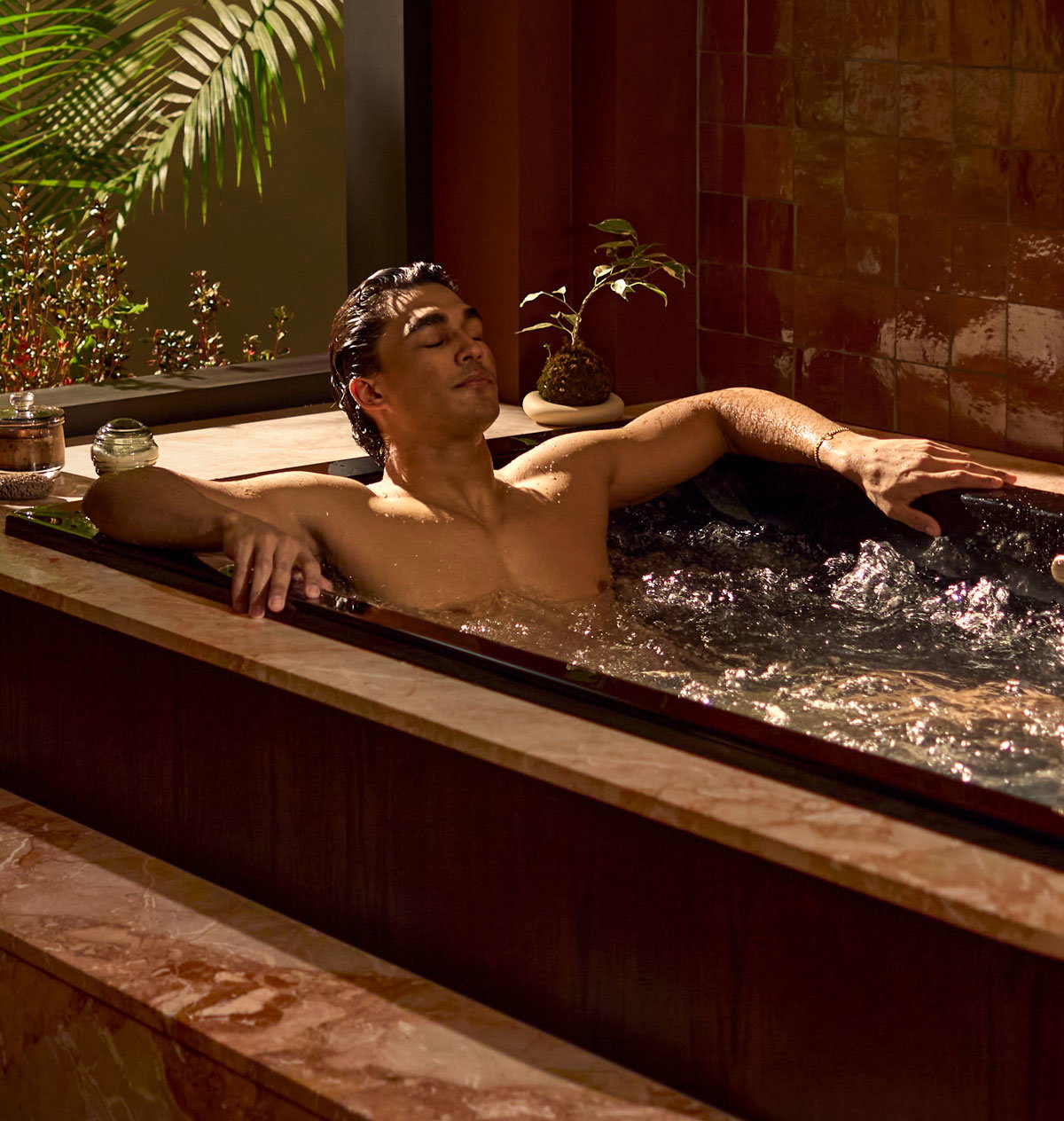 Person relaxing in a bathtub with hydrotherapy jets beside a large window.
