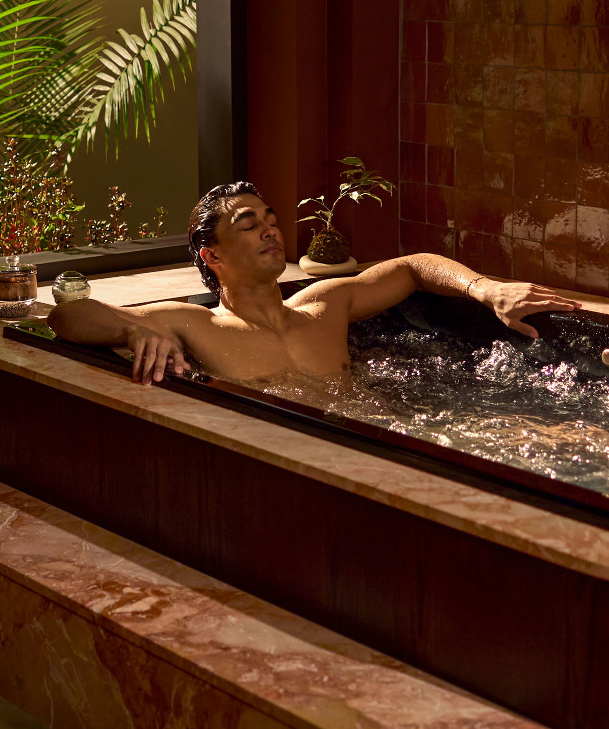 Person relaxing in a bathtub with hydrotherapy jets beside a large window.