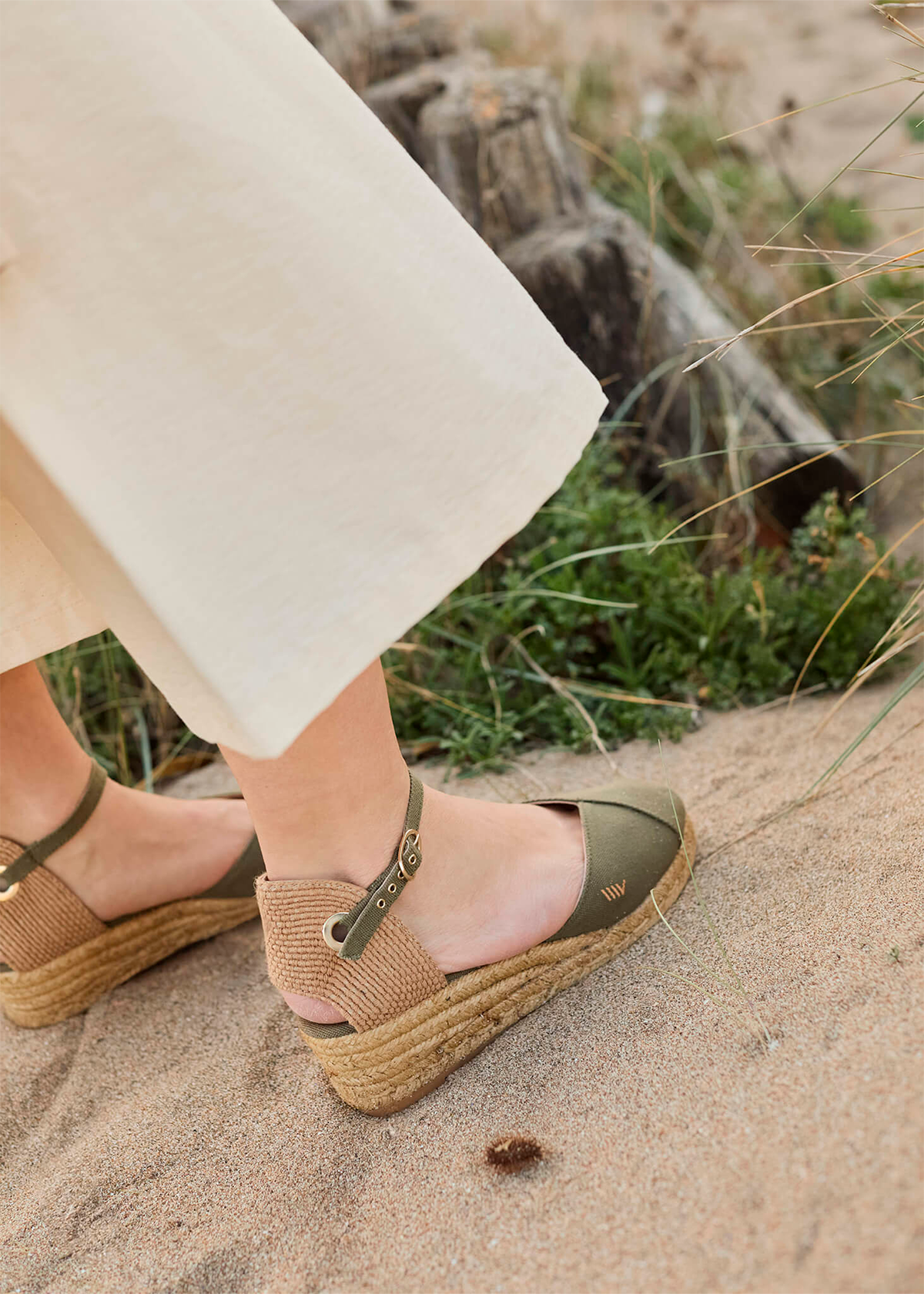 Close-up of olive Pubol canvas ankle-strap wedge espadrilles by Viscata worn on a sandy coastal path.